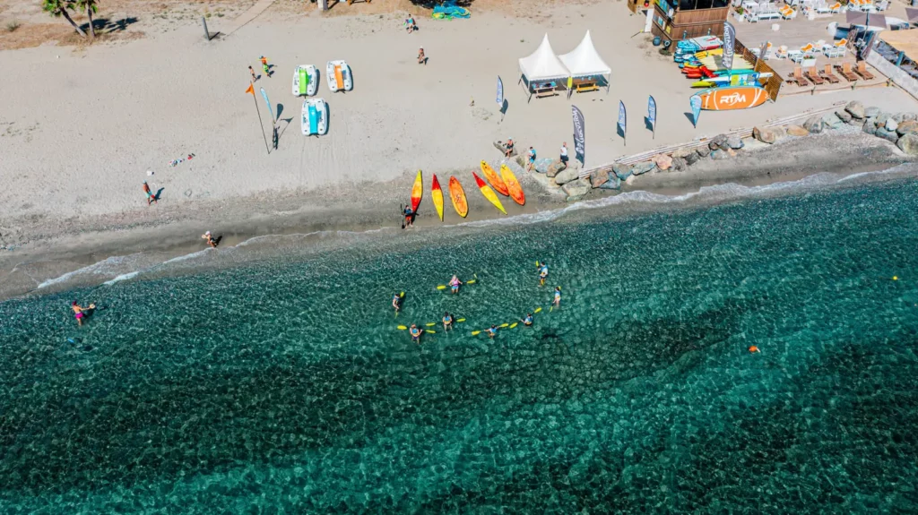 Kayak en Corse - activités nautiques à merendella Vue aérienne d’une plage corse avec kayaks, paddles et groupe d’enfants pratiquant une activité nautique dans une eau transparente.