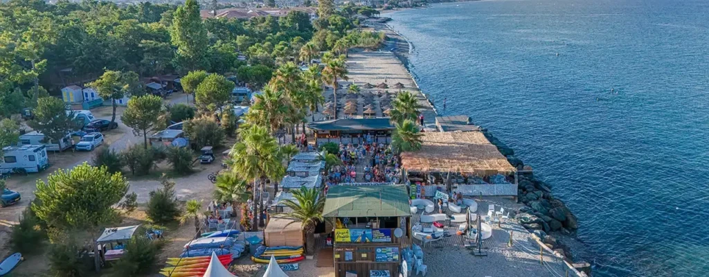 camping-corse-bord-de-mer-luxe-merendella-1 Centre de loisirs et restaurant de plage ombragé par des palmiers dans un camping de luxe en Corse, avec de nombreux clients et des kayaks colorés.