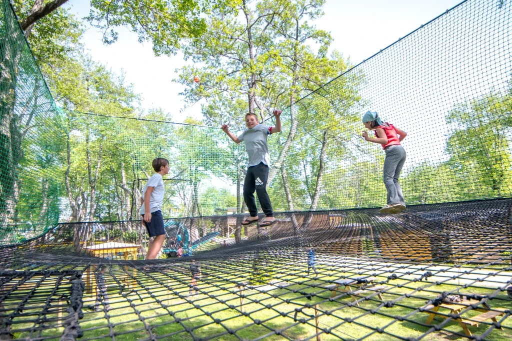 accrobranche-parcours-kid-camping-merendella Enfants qui sautent et s'amusent sur un grand filet de trampoline suspendu en plein air dans un parc de loisirs en Corse.