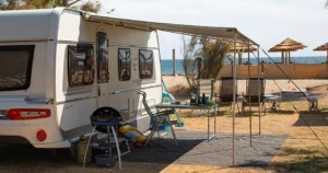 Caravane moderne avec auvent déployé, salon de jardin et barbecue installés sur un emplacement de camping en front de mer.
