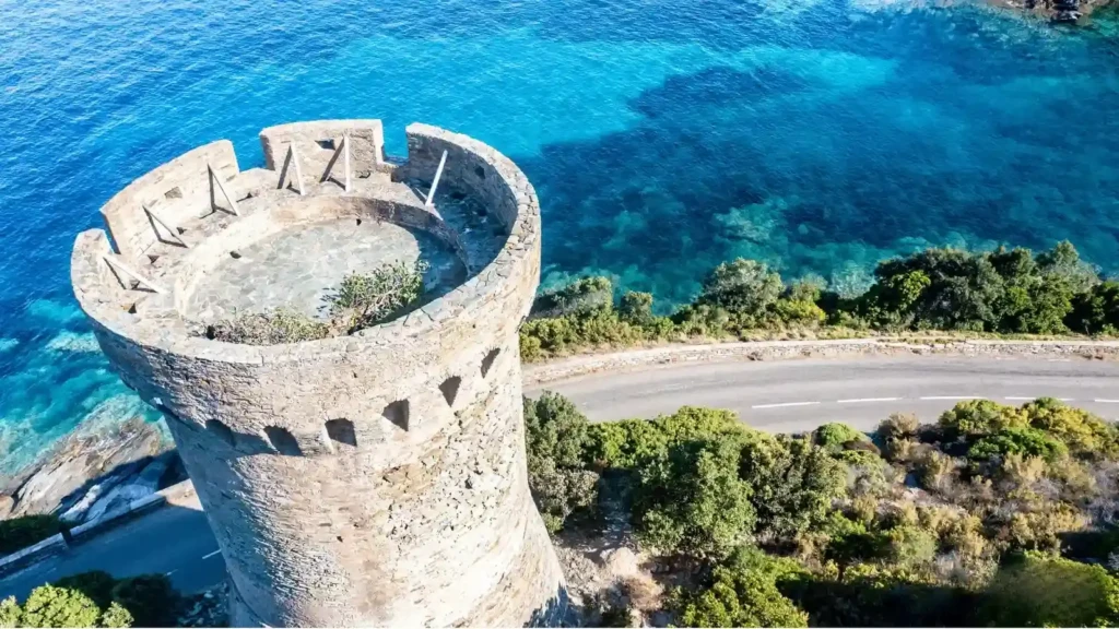 patrimoine-corse-tour-genoise-littoral-1 Vue aérienne d'une tour génoise historique en pierre surplombant les eaux turquoise de la Méditerranée le long de la côte corse.